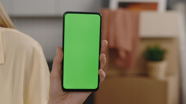 Close-up of female hand holding green chroma key screen smartphone indoors in moving home blurred background. Gadgets and moving concept.