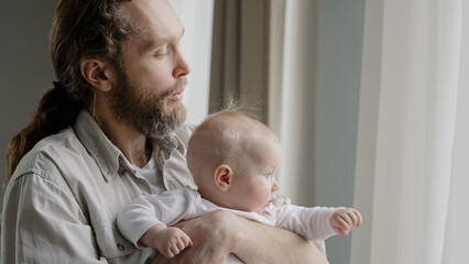 Close-up caucasian father bearded daddy holding baby daughter son infant newborn looking out window with curtains dad blowing to short blonde hair of kid child playing at home. Fatherhood protection