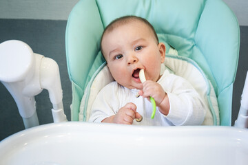 A hungry baby is gnawing on a plastic spoon at the table on a high chair. Teething, whims, itchy gums, introduction of complementary foods