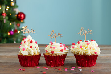 Three cupcakes decorated on blue background with LOVE sign and out-of-focus Christmas tree in background. Copy space.