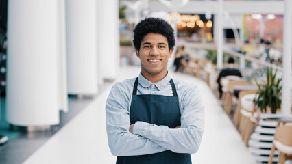 Confident African 30s restaurant worker biracial guy strong position standing indoors barman seller waiter in apron looking to camera successful cafe owner posing with crossed arms small business - Powered by Adobe