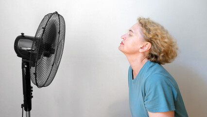 .middle aged blond woman  enjoying air flow from electric fan at home, Happy woman with big fan