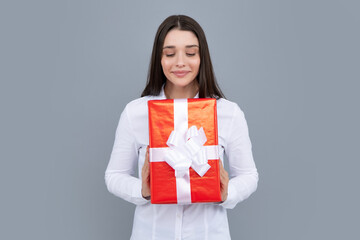 Fototapeta premium Surprised woman holding gift. Smiling beautiful young brunette woman in shirt hold present box with gift ribbon bow isolated on bright gray background, studio portrait. Birthday concept.