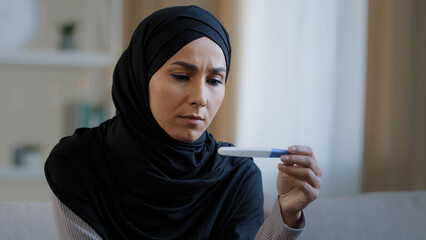 Shocked unhappy islamic young woman arabian muslim girl in hijab female adult pregnant feeling fear looking at positive pregnancy test result sit at home worrying about unwanted children bad reaction