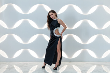 Obraz premium Beautiful caucasian brunette-haired woman wearing black evening gown with a slip stands by white illumonated wall. Full length portrait. People theme.