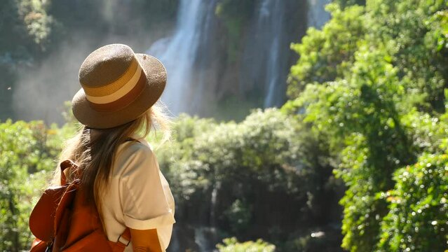 Travel woman enjoy amazing view of waterfall nature. Female traveler in hat with backpack in Asia adventure or journey. Cinematic video of girl in tropical vacation.