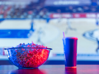 A sports match on the screen of a large plasma TV. Popcorn in a glass bowl and a drink in a plastic...