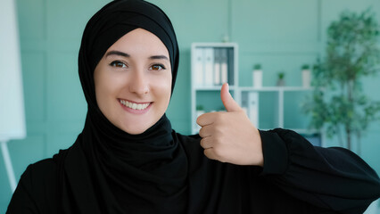 Close-up female portrait in office Muslim happy satisfied contented Islamic Arabian woman wearing black hijab looking at camera smiling toothy girl manager showing like thumbs up good perfect results
