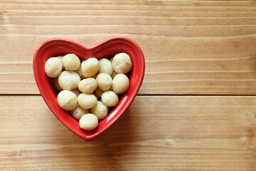 Macadamia nuts lie in a heart-shaped ceramic bowl, which stands on a wooden table. View from above