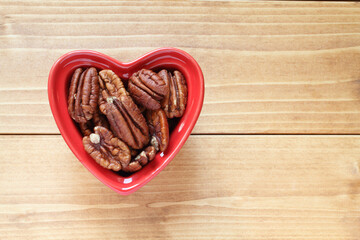 Pecans lie in a heart-shaped ceramic bowl, which stands on a wooden table. View from above