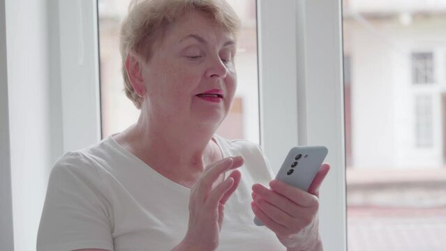 Older Middle Aged Blond Woman using and text messaging by mobile phone near Window with old town background, indoor, Relaxed mature old 60s woman, search ecommerce offers on cell phone technology devi