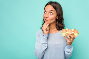 Young hispanic woman holding eggs isolated on blue background looking sideways with doubtful and skeptical expression.