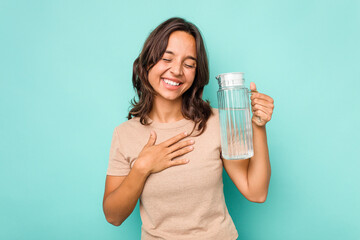 Young hispanic woman holding a water of jar isolated on blue background laughs out loudly keeping hand on chest.