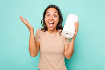 Young hispanic woman holding kitchen roll isolated on blue background receiving a pleasant surprise, excited and raising hands.