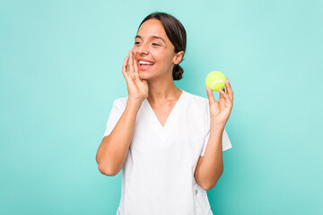 Young hispanic physiotherapy holding a tennis ball isolated on blue background shouting and holding palm near opened mouth.