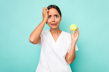 Young hispanic physiotherapy holding a tennis ball isolated on blue background being shocked, she...