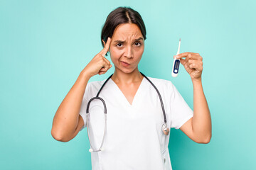 Young hispanic nurse woman holding a thermometer isolated on blue background pointing temple with finger, thinking, focused on a task.