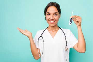 Young hispanic nurse woman holding a thermometer isolated on blue background showing a copy space on a palm and holding another hand on waist.