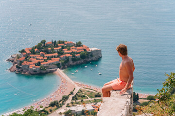 Obraz premium a man poses on a mountain with a view from above of the city of Sveti Stefan in Montenegro, a picturesque bay