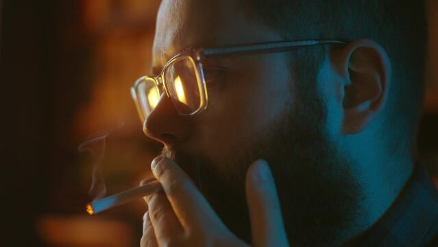 Pensive bearded man in eyeglasses lighting cigarette, inhaling smoke, thinking