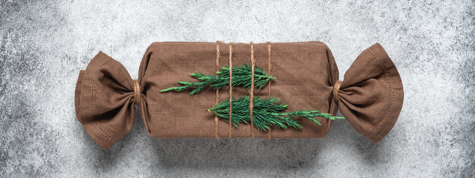 Christmas gift wrapped in fabric with coniferous green branches, gray concrete background. Gift in furoshiki style. Zero waste concept. Top view, flat lay. Banner. Selective focus.