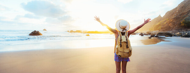 Happy female traveller with backpack and hands up enjoying sunset on the beach - Delightful woman...