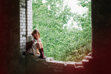 Fototapeta premium Art work on the theme of the war in Ukraine. A frustrated Ukrainian woman in national dress sits in the window frame of a building bombed by the Russian army.