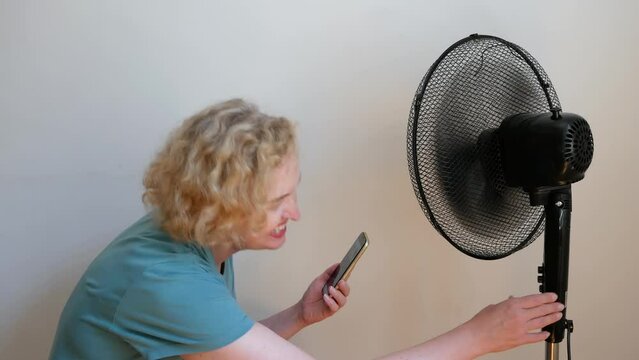 middle aged blond woman  with mobile phone enjoying air flow from electric fan at home, remote control of electic fan