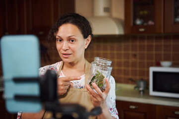 Charming African American woman, successful food blogger, pleasant housewife, showing a jar with culinary herbs to the camera on her smartphone, on a tripod while recording video content for her blog