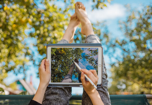 Moscow, Russia - August 17, 2022 : Girl sits in the garden on swing upside down and draws on iPad Pro sketches with her legs on background of the sky. Inspiration, creativity, self-development, design