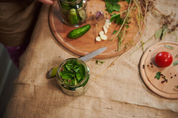 View from above of a glass jar for canning with stacked cucumbers and poured marinade and a wooden board with chopped ingredients, garlic an fresh culinary herbs on a linen tablecloth. Still life