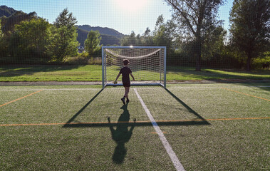 Fototapeta premium Young boy playing soccer on the football field at sunset, shooting at empty goal posts