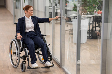 Red-haired caucasian woman in a wheelchair trying to open the door in the office. 