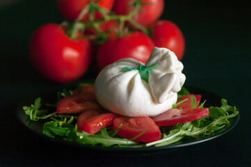 on a black plate, spinach salad and arugula greens, sliced tomatoes and Italian burrata cheese on a black background, a bunch of red tomatoes is visible in the background, side view.