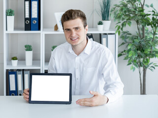 Online presentation. Computer mockup. Digital life. Smiling office man pointing to tablet computer with blank screen sitting desk in light room interior.