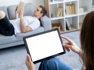 Online education. Computer mockup. Home routine. Unrecognizable woman holding tablet computer with blank screen while defocused man reading book in light room interior.