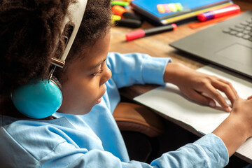 African-American girl doing homework,making notes,using headphones and laptop at home.Back to school concept.School distance education at home,home schooling,e-learning,diverse people.