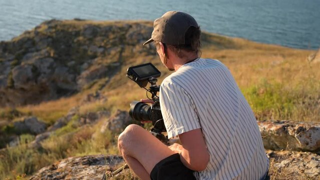 Filmmaker with camera on tripod changes settings and shoots a sea sunset.