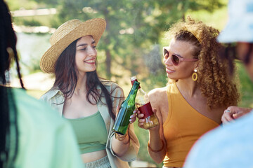 Portrait of two smiling young women clinking beer bottles outdoors while enjoying party with...