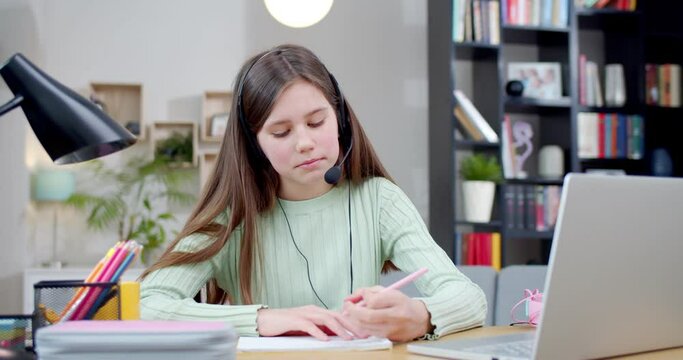 Caucasian pretty teen girl in headset with mic and headphones videochatting online on laptop computer and having coaching class. At home. Schoolgirl learning by video schooling class with video call.