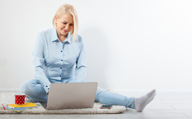 Happy middle aged woman sitting relaxed on the floor using laptop for entertainment. The concept of leisure and work with a cup of coffee at home.