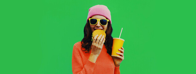 Portrait of stylish young woman drinking juice with burger fast food on green colorful background