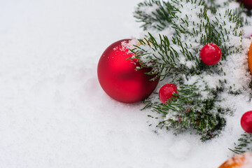 Close-up photo of a large red Christmas toy in the form of a ball and several small ones on a branch of a coniferous tree lying in the snow. Left space for text