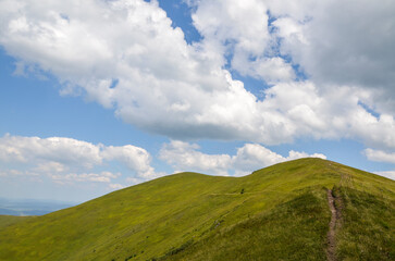 Hiking trail through mountain ridge with green grassy slopes under blue sky with clouds on summer day. Carpathian Mountains, Ukraine