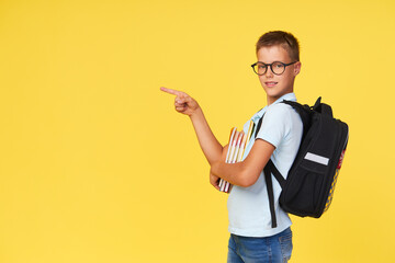 Portrait of a schoolboy in glasses with textbooks and a backpack on a yellow background pointing to...