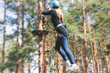 Fototapeta premium View of high ropes course, process of climbing in amusement acitivity rope park, passing obstacles and zip line on heights in climbing safety equipment gear between the trees, summer sunny day
