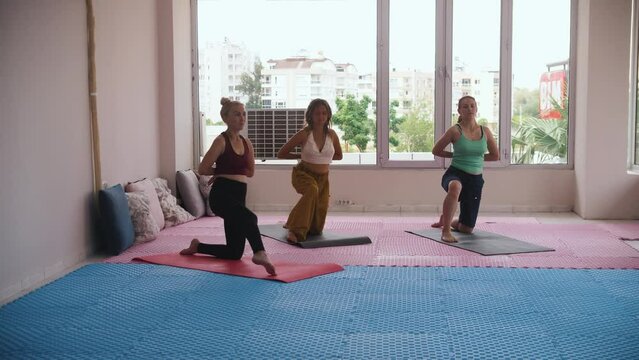 Three women standing on yoga mats on their knees and doing the exercises in the studio with big windows