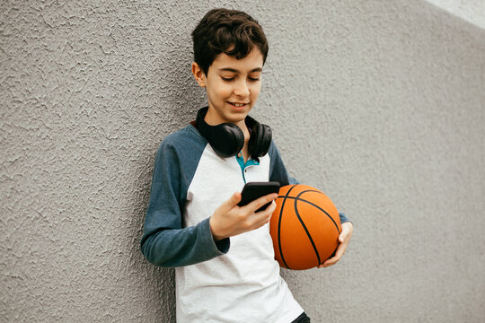 Teen boy using smartphone after playing basketball outdoors.
