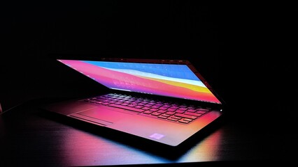 Laptop with half lid open on a table lit with colorful desktop screen wallpaper in a dark room	
