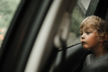 Thoughtful little blond baby boy seated in car backseat looking through rain covered window in...